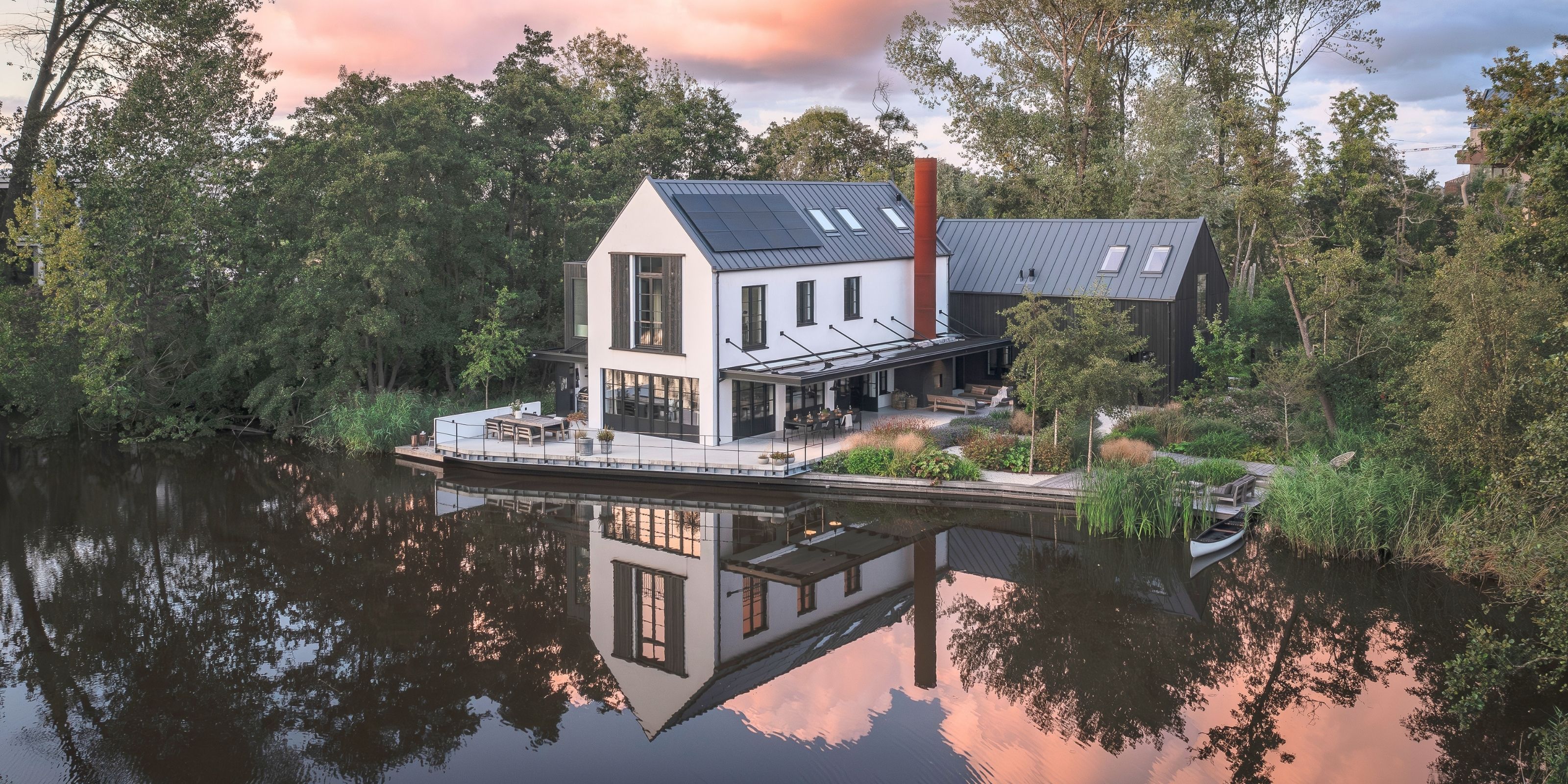 The Factory House at golden hour: the white house with its red chimney and the black timber barn beside it, mirrored in the still water under a pink sky.