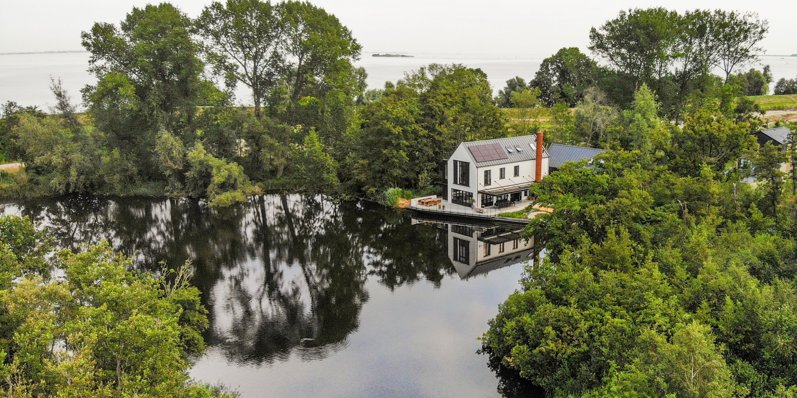 Aerial view of The Factory House on the water, surrounded by trees, with the IJmeer in the distance.