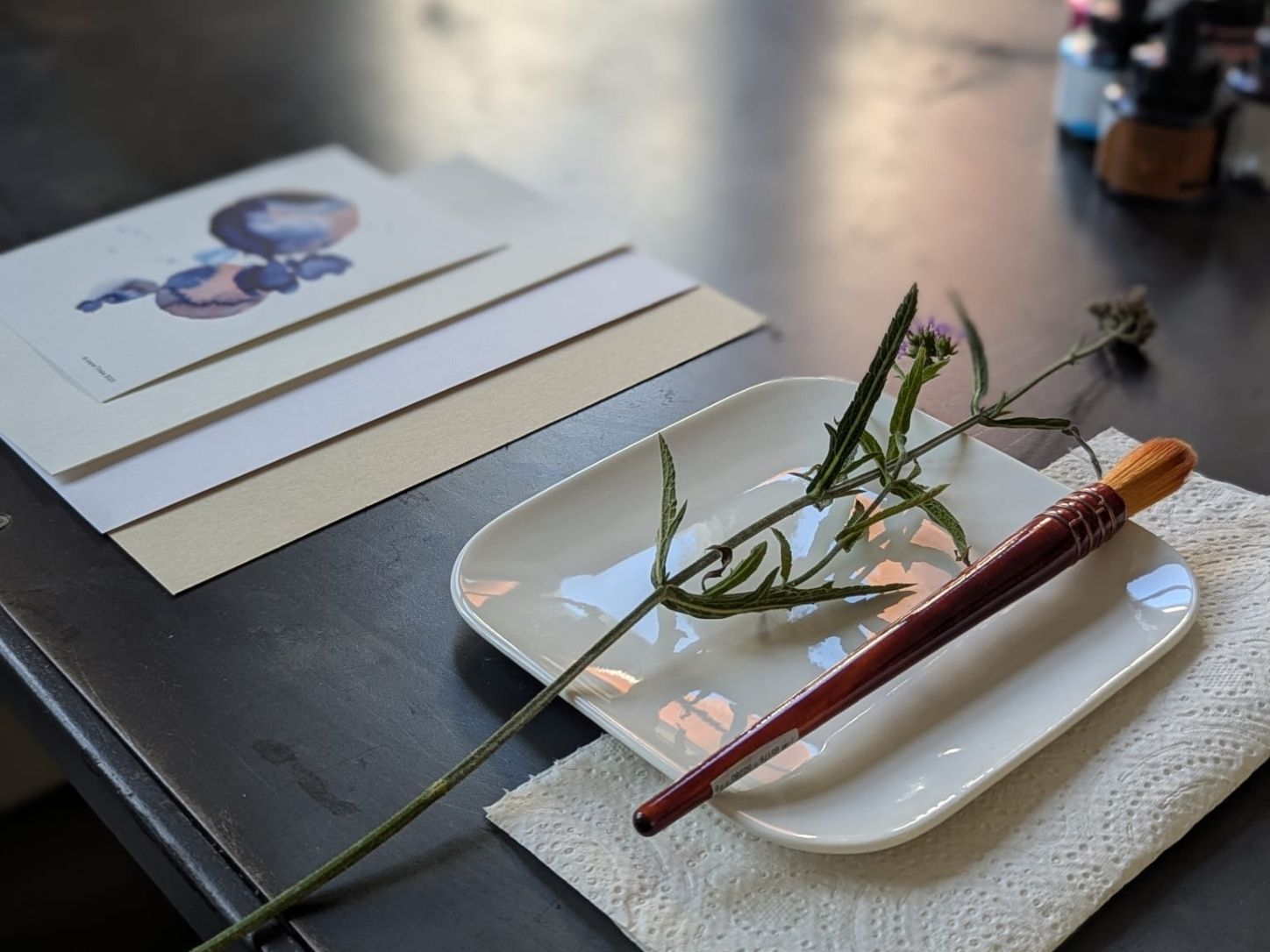 Workshop detail: an ink painting on paper, a brush and a flower stem on a white dish.