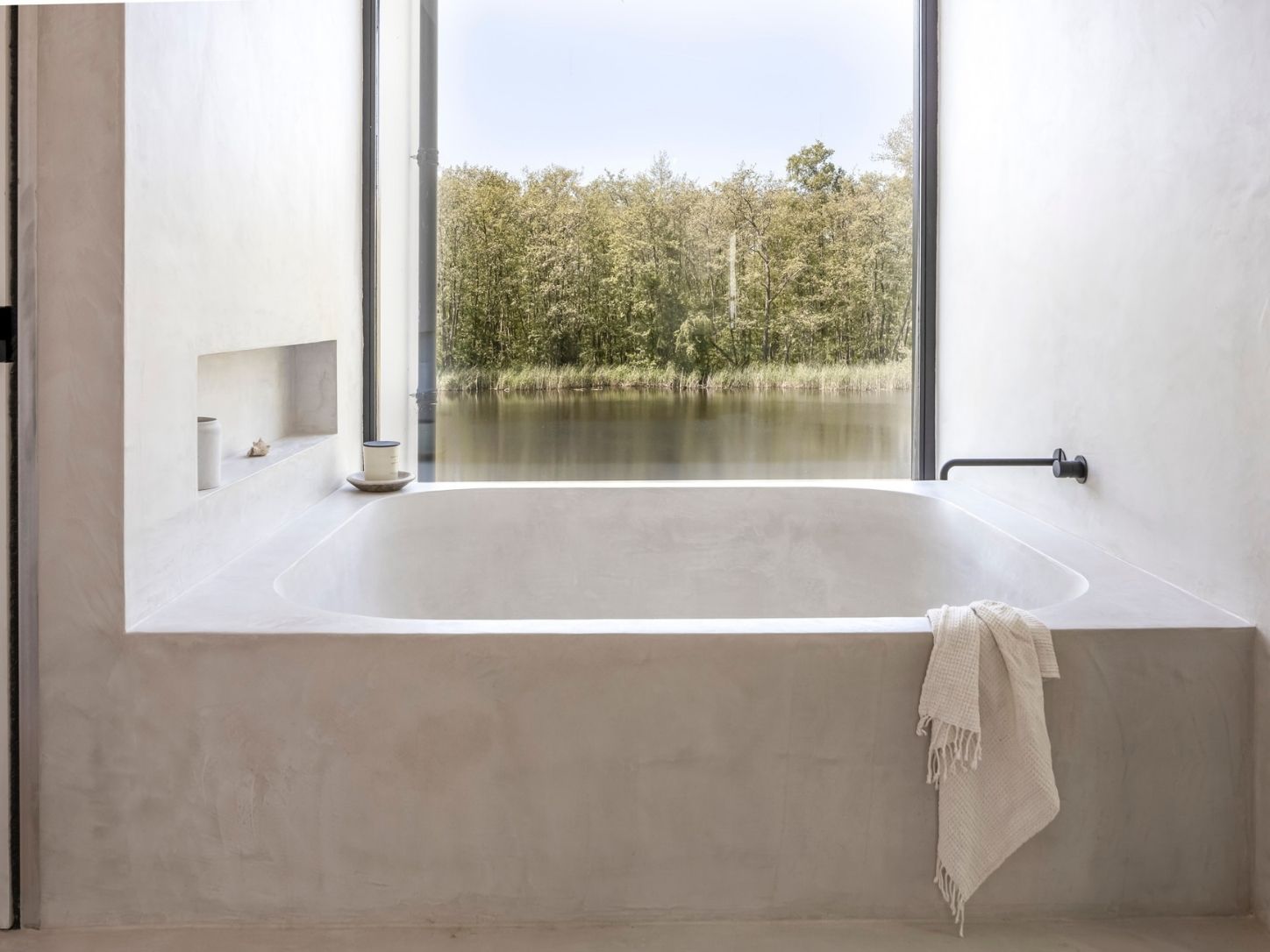 The microcement bath in front of a tall window, looking out over the water and the forest, with a linen towel over the edge.
