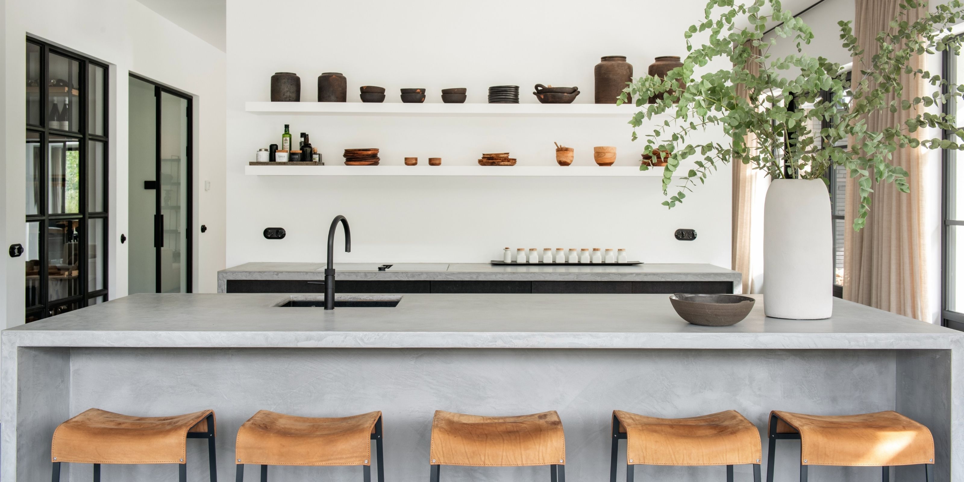 The microcement kitchen island with five leather bar stools, and open shelves with earthenware behind it.