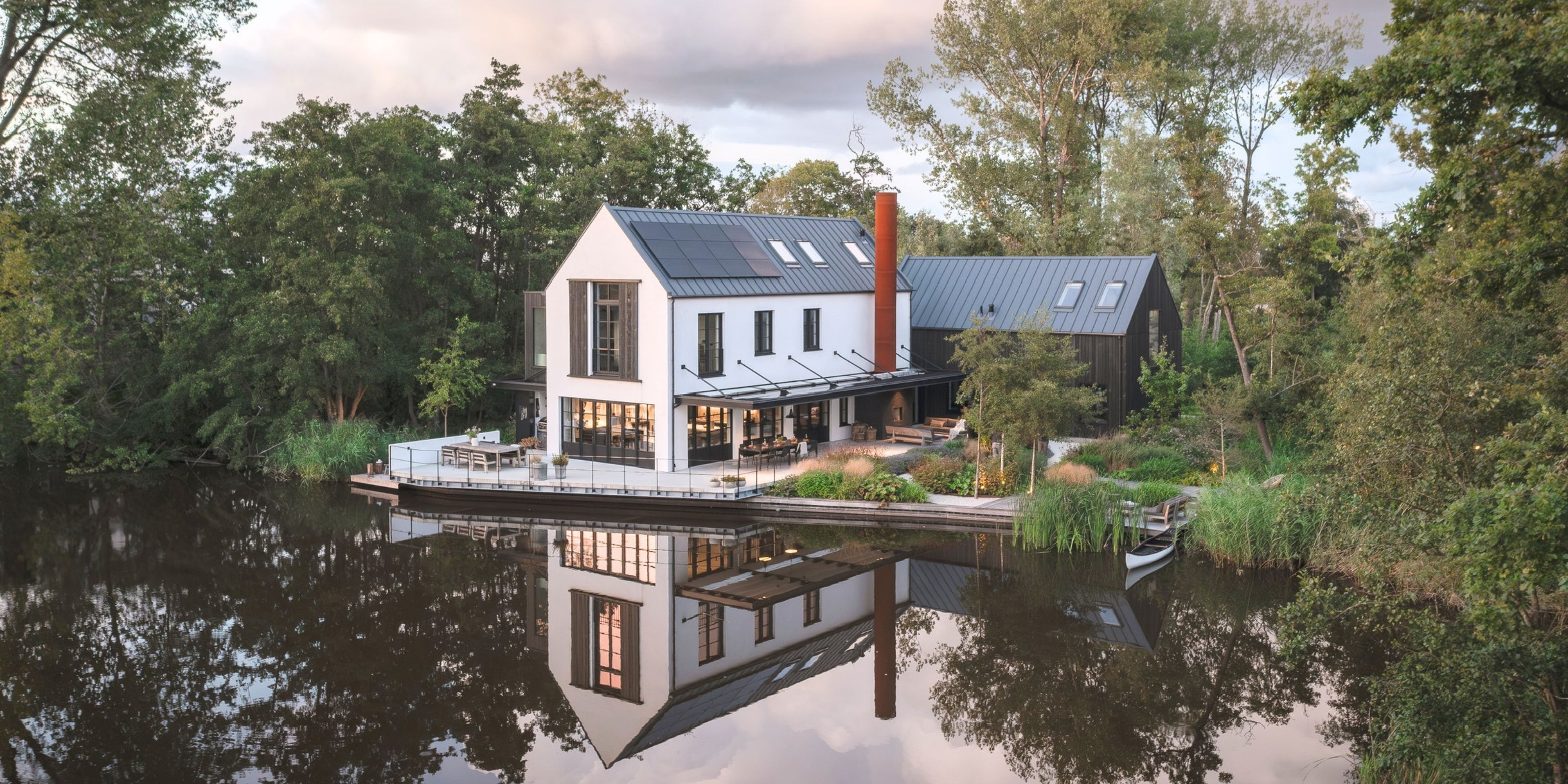 The house and the Shou Sugi Ban barn at dusk, with the terrace at the water's edge and the house reflected in the water.