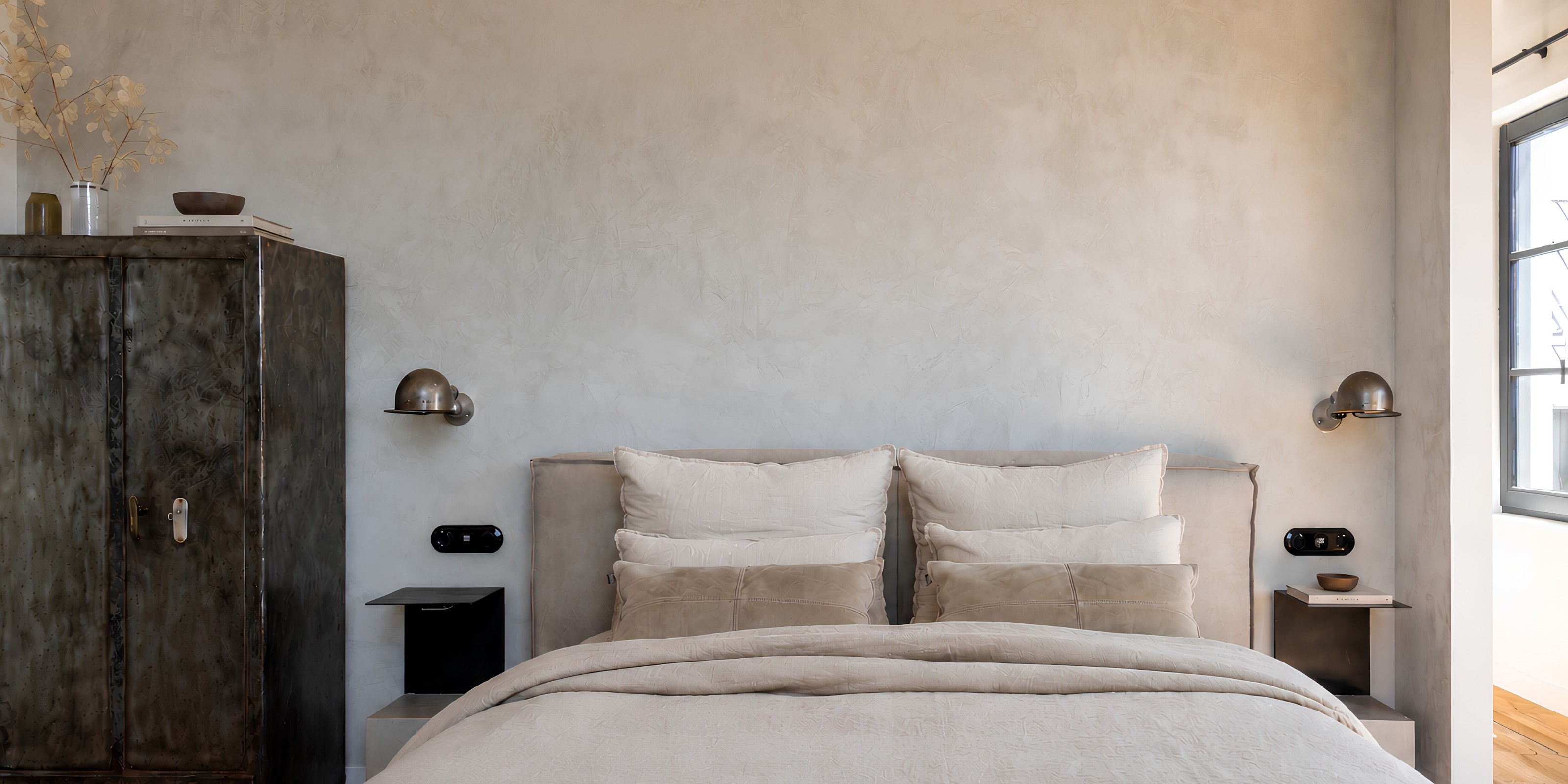 The bedroom: a bed with linen cushions against a textured plaster wall, two steel wall lights, a dark metal wardrobe on the left and a window on the right.