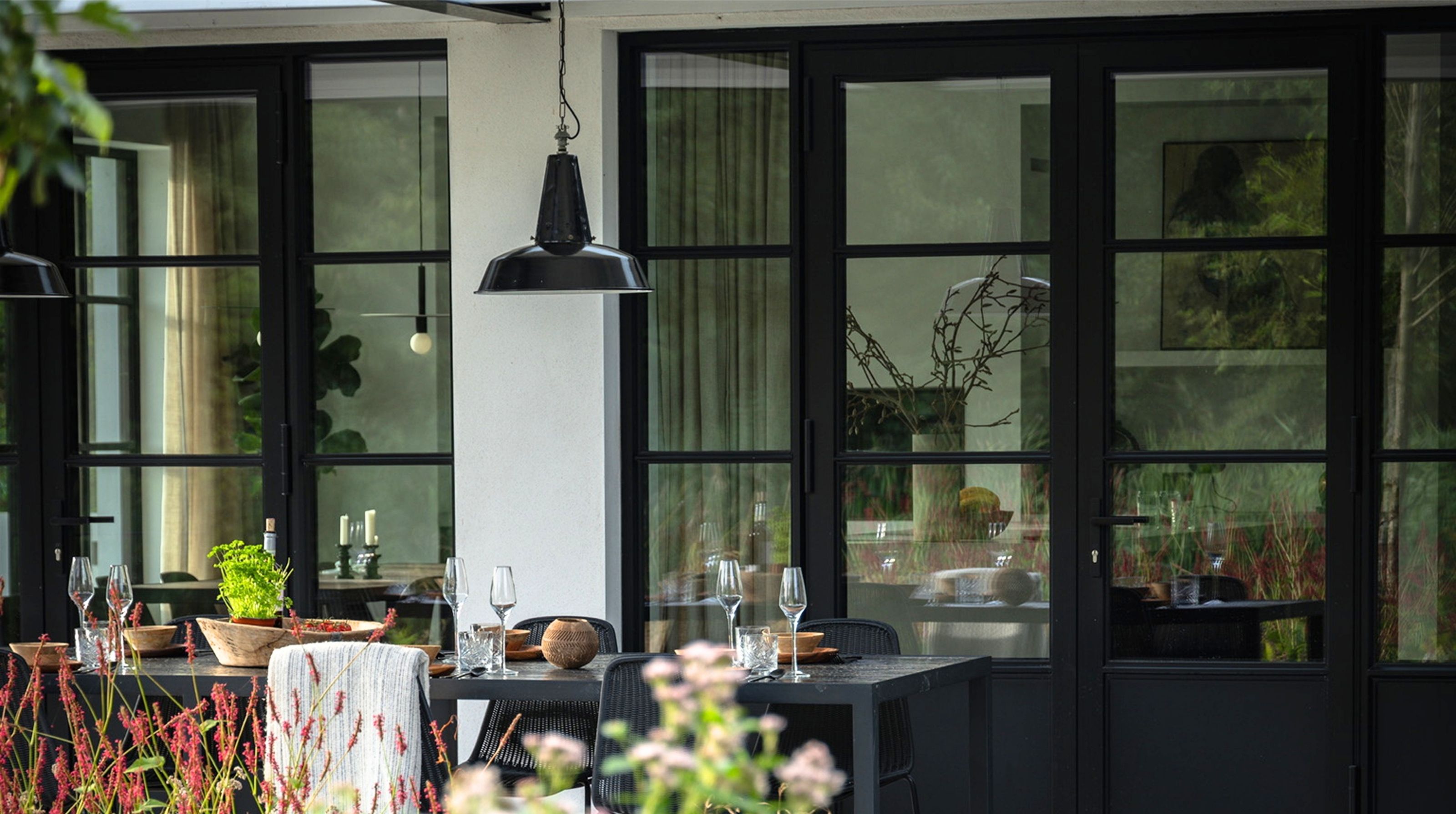 The terrace laid for dinner in front of the black steel doors, with flowering plants in the foreground.