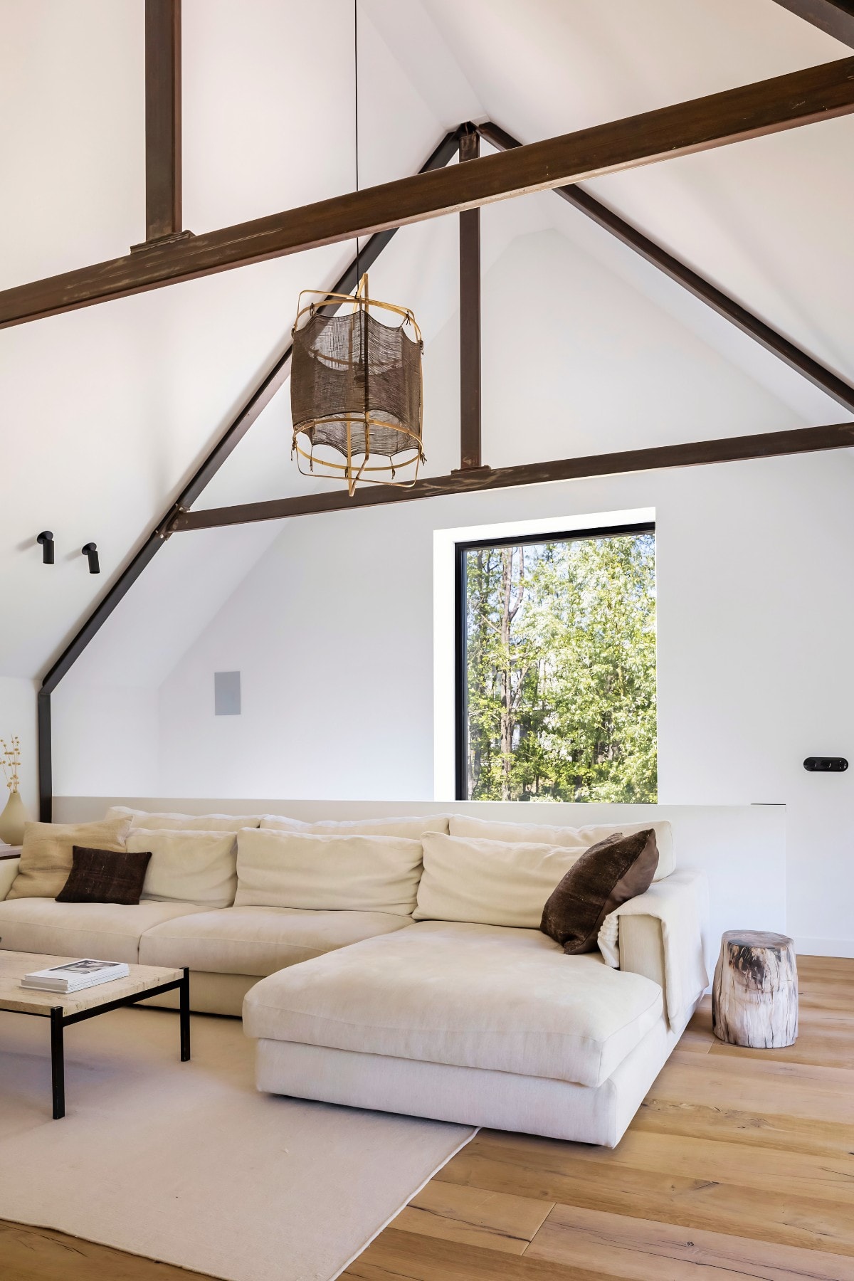 The second living room: a large cream sofa under a high pitched ceiling with dark steel beams, a woven pendant light, and a tall window onto the forest.