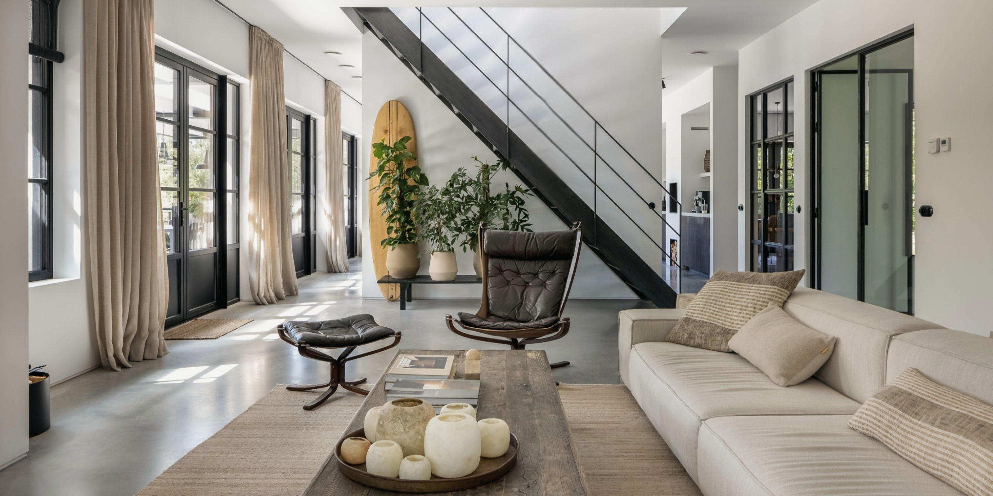 The living room: tall steel windows with linen curtains, a concrete floor, the steel staircase and a surfboard against the wall.